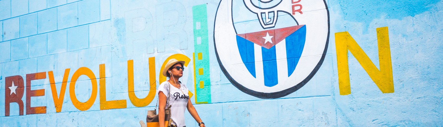 A photograph of a woman walking on the street in front of a Socialist propaganda mural in cienfuegos, cuba